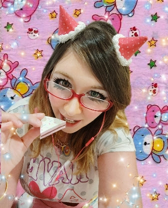 Beautiful person with glasses and a cake in front of a colorful, cartoon-themed background, wearing devil horn hair clips.